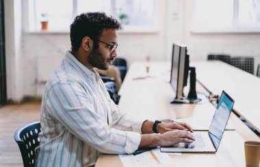 Latin kökenli sakallı erkek işçilerin günlük kıyafetleri ve gözlükleriyle masada dizüstü bilgisayarları ve kahve bardaklarıyla oturup ofiste yazı yazarken ve çalışırken