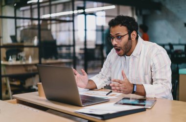 Yoğunlaşmış sakallı, günlük kıyafetlerle serbest çalışan, dizüstü bilgisayarlı ve not defterli, yaratıcı çalışma alanı projesinde çalışırken ağzı açık bir şekilde hayrete düşmüş bir erkek.