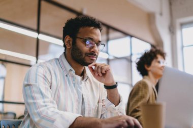 Düşük açılı, sıradan kıyafetler ve gözlüklü İspanyol bir erkek çalışanın ofiste bulanık arka planda çalışırken bir fincan kahveyle masada oturması ve çenesine dokunması.