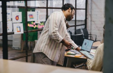Dizüstü bilgisayarı incelerken ve yaratıcı çalışma ortamında masada oturan ekin işçisiyle çalışırken günlük giysiler içindeki konsantre sakallı erkek mimarın yan görünüşü