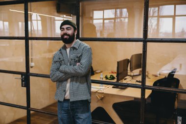 Pozitif, genç, sakallı, günlük giysiler içinde, kollarını kavuşturmuş, cam duvarın yanında durmuş, kameraya bakıyor ve yaratıcı çalışma alanında çalışıyor.