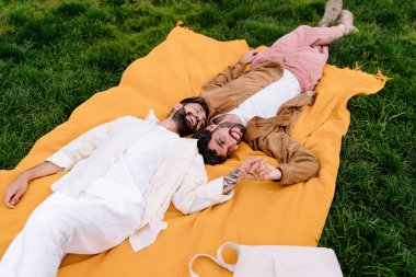 Şehir parkında battaniyenin üzerinde uzanan ve romantik bir randevu sırasında el ele tutuşup eğlenen sevgili çiftlerin kıyafetlerinden.