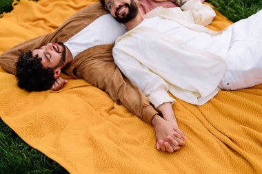 Sarı battaniyenin üzerinde uzanan ve gündüz vakti parkta rahatlarken ve el ele tutuşurken birbirini okşayan gündelik giysiler içindeki aşk dolu genç çiftten.