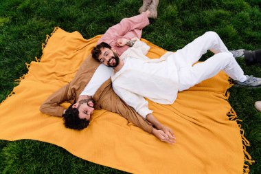 Sakallı, genç, eşcinsel bir çiftin battaniyeye uzanmış, gözlerini kaçırırken, erkek gözlüklü, bacak bacak bacak üstüne atmış ve birbirlerinin ellerine dokunurken görüntüsü.