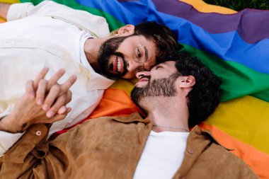 Mutlu, genç, sakallı, sıradan giyinmiş İspanyol bir çiftin güneş ışığında LGBT bayrağının alınlarına dokunup el ele tutuşurken birbirlerine bakışlarının üst görüntüsü.