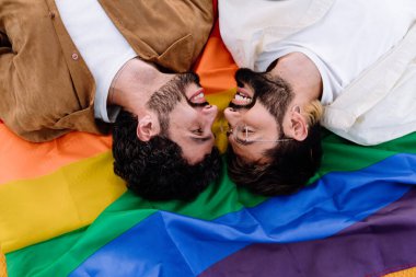 Gülümseyen genç, sakallı İspanyol eşcinsel çiftin, LGBT bayrağında uzanıp gün ışığında dinlenirken, birbirlerine baktıkları en güzel manzara.