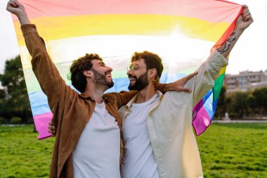 Gökkuşağı LGBT bayrağını tutan mutlu genç erkek çift çayırların bulanık arka planında dikilirken birbirlerine bakıyorlar.