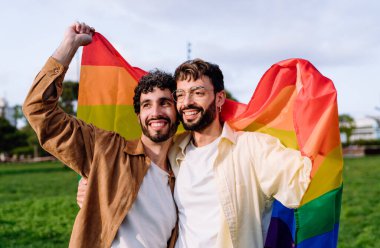 Pozitif İspanyol siyah saçlı eşcinsel çift gökkuşağı bayrağıyla sarılıyorlar ve güneşli bir günde yeşil çimlerin üzerinde dikilirken başlarını çeviriyorlar.
