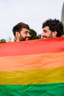 Hafta sonları parkta gökkuşağına karşı gökkuşağı bayrağı tutarken gülümseyen ve birbirlerine bakan koyu saçlı neşeli genç gay erkeklerin yan görüntüsü