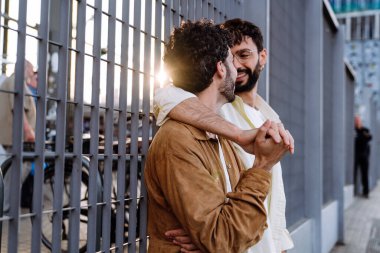 Siyah saçlı, sakallı, genç eşcinsel bir çiftin gündüz birbirlerine bakarken metal çitlerin etrafında dikilmesi.