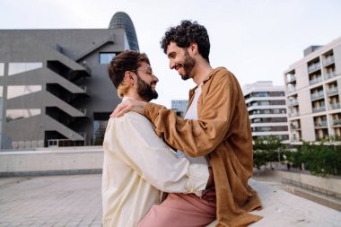 Romantik bir buluşma sırasında şehrin çatısında kucaklaşırken gülen ve birbirlerine bakan gündelik kıyafetli İspanyol eşcinsel çiftin yan görüntüsü.