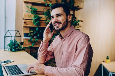 Telefon görüşmesi sırasında dizüstü bilgisayar üzerinden uzak iş sahip yakışıklı adam gülümsüyor, mobil bağlantı arkadaşıyla kafede vakit cep üzerinde konuşurken neşeli hipster adam memnun