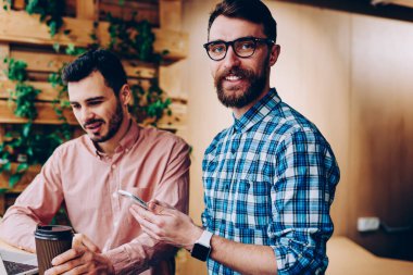 Neşeli bir erkek, kameraya bakıyor ve telefonu tutuyor. Arkadaşı da bilgisayardan internetten bilgi okuyor. Refah içindeki bir çalışan, projedeki meslektaşıyla iş birliği yapıyor.