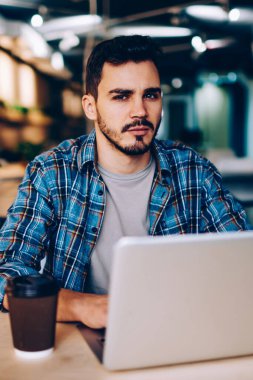 Üniversitede internet ve kablosuz internet kullanarak sınav hazırlığı yapan ciddi bir hipster çocuğun portresi. Kameraya bakan genç adam, birlikte çalıştığı yerdeki dizüstü bilgisayarları kullanarak hayal kırıklığına uğradı.