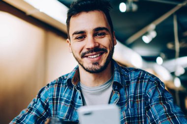 Takipçilerle sohbet etmek için telefonu kullanan ve sosyal medyadan zevk alan yeni arkadaşları onaylayan neşeli erkek blog yazarının portresi kahve molasında kameraya bakıp gülümseyen öğrenci