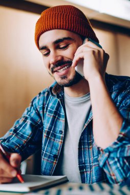 Neşeli erkek yaratıcı tasarımcı defterine çizim yapıyor telefon konuşması yapıyor, gülümseyen hipster adam üniversite kütüphanesinde otururken fikir alışverişi yaptığı için arkadaşına telefon ediyor.