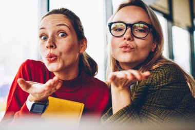 Yarım uzunluk portre neşeli genç kadınların zamana Cafe iç oturma sırasında kameraya hava öpücük gönderme rahat kıyafet giymiş. Poz ve having fun olumlu ve büyüleyici iki arkadaşları