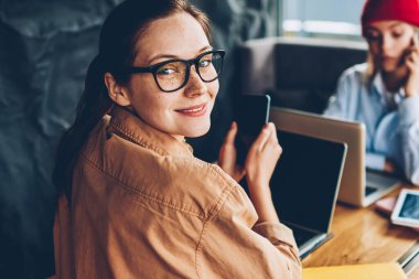 Gözlük vizyon düzeltme posta Smartphone cep telefonu ile coworking ve birlikte eğitim hipster arkadaşıyla oturan ağlarda denetlerken kameraya gülümseyen neşeli genç kadın portresi