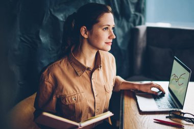 Kitap okuyan ile düşünceli zeki hipster öğrenci dizüstü cihaz oturan ve 4 g kullanarak grafik düzenleri hakkında çalışma elinde kablosuz internet. Modern netbook çalışan genç kadın durulması