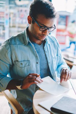 Serin Eyewear tahta masada oturan kafeteryada çalışan yakışıklı zeki hindu Tasarımcısı resim kırpılmış. Kalem ve kağıt kapalı kullanarak yeni planları oluşturma yetenekli erkek mimar