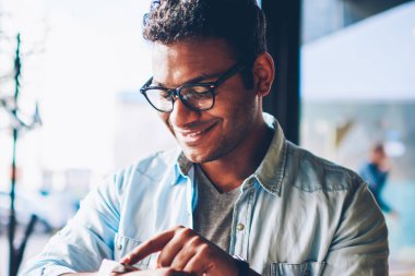 İyi görünümlü gülümseyen hindu işadamı havalı gözlükler dijital smartwatch kapalı oturma yapılandırma içinde kırpılmış görüntü. Olgun yakışıklı mutlu erkek kişi vaktinizi kafede modern cihaz ile