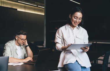 Görme düzeltmesi için klasik göz giysisi kullanan mutlu Asyalı çalışan uygulama üzerinden web sitesinde bilgi taramak için dijital tablet kullanıyor, çevrimiçi rezervasyon yapan gözlüklerde başarılı grafik tasarımcısı
