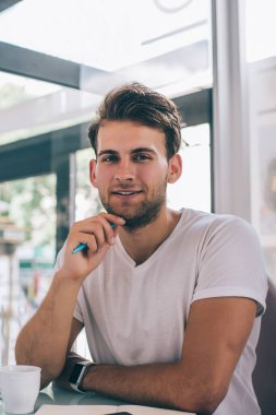 Tişörtlü neşeli genç yakışıklı adam kameraya bakıyor ve arkadaşı beklerken çenesine kalemle dokunuyor ve kafede oturuyor.