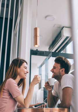 Düşük açılı gülen adam ve kadın gündelik giysiler içinde birbirlerine bakıyorlar. Masada oturmuş kahve fincanları tutuyorlar ve modern ofiste sohbet ediyorlar.