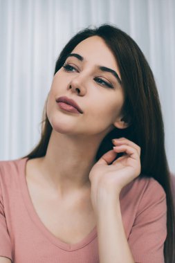 Pembe tişörtlü çekici esmer rüya gibi bir yüz ifadesiyle başka tarafa bakıyor ve arka planda beyaz pencere perdeleriyle el kaldırıyor.