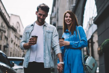 Aşağıdaki yakışıklı, memnun, sıradan kıyafetler giymiş, elinde içecekle kız arkadaşıyla el ele tutuşup gülen ve şehir merkezinde cadde boyunca yürüyen adamdan.