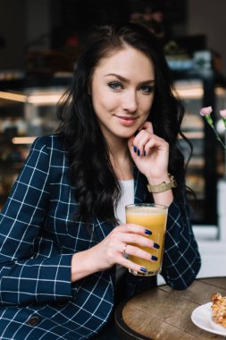 Resmi giysiler içinde neşeli bir kadın yuvarlak masada oturuyor ve sabah kafede kahvaltı ederken taze meyve suyu içiyor.