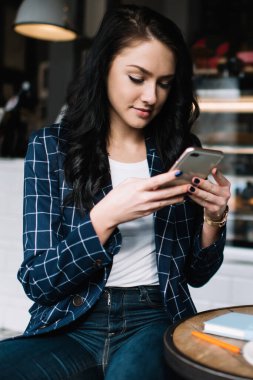 Çağdaş kafe projesinde çalışırken öğle yemeği sırasında iş yerinin fotoğrafını çekmek için günlük kıyafetlerini giyen rahat bir kadın telefon kullanıyor.