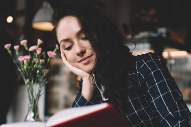 Moda kıyafetli, rahat bir kafedeki ahşap masada oturan ve kahve molası sırasında ilginç kitaplar okuyan bir kadın.