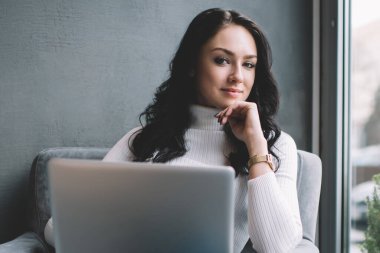Düşünceli kadın serbest çalışan, pencerenin yanında otururken kameraya sırıtarak bakıyor ve gündüzleri yeni iş planı üzerinde çalışıyor.