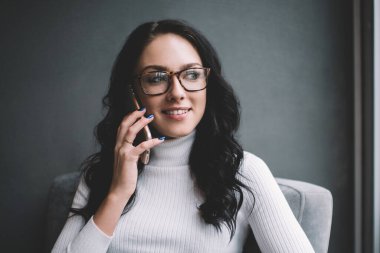 Modern kafede rahat bir koltukta otururken cep telefonuyla konuşan ve kahve içen genç bir kadın.