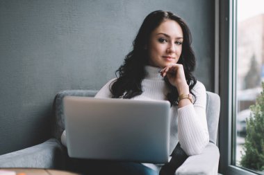Düşünceli kadın serbest çalışan, pencerenin yanında otururken kameraya sırıtarak bakıyor ve gündüzleri yeni iş planı üzerinde çalışıyor.
