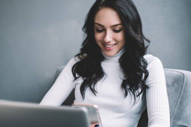 Beyaz süveter giyen, rahat koltukta oturan ve dizüstü bilgisayar projesinde çalışırken akıllı telefondan konuşan pozitif kadın serbest yazar.