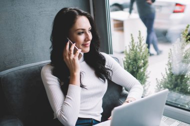 Kafede dizüstü bilgisayarla çalışırken koltukta oturup akıllı telefondan konuşan serbest çalışan düşünceli bir kadın.