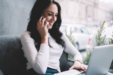 İyimser kadın, beyaz kazaklı, rahat koltukta dinlenirken ve cep telefonuyla sohbet ederken gülümseyen ve bilgisayarını karıştıran serbest yazar.