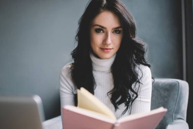 İçerikli, rahat giyimli genç bir bayan, evdeki modern oturma odasında dizüstü bilgisayarla koltukta oturup kitap okurken kameraya bakıyor.