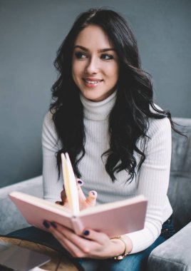 Sıradan giyinen mutlu bayan öğrenci kitap sayfalarını çevirirken koltuğuna oturur ve başka tarafa bakar ve evdeki modern oturma odasında rapor hazırlamak için dizüstü bilgisayar kullanır.