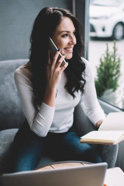 Gülümseyen kadın serbest yazar, yaratıcı çalışma alanında oturuyor ve elinde dizüstü bilgisayarla cep telefonuyla konuşuyor.