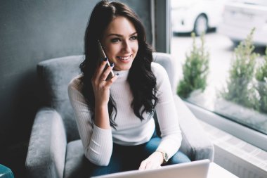 Dizüstü bilgisayar kullanan ve cep telefonuyla konuşan mutlu kadın serbest yazar yaratıcı iş yerinde oturup iş projesinde çalışıyor.