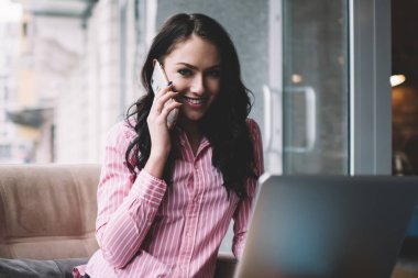 Modern kafeteryadaki rahat koltukta rahat bir şekilde cep telefonuyla konuşurken dizüstü bilgisayar ve not defteriyle oturan serbest çalışan genç bir kadın.