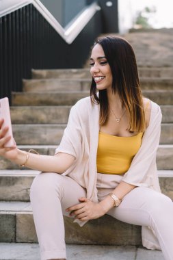 Neşeli, sıradan giyinmiş esmer kadın yaz günü cep telefonunda selfie çekerken merdivenlerde oturuyor ve gülümsüyor.
