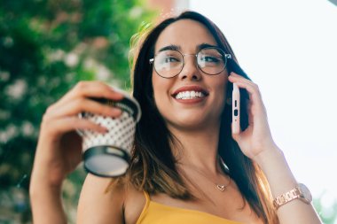 Gözlüklü, yaz tişörtlü, tek kullanımlık kahve bardağı tutan, geniş pencereli ve gözlerini kaçıran konuşkan genç bir kadın.