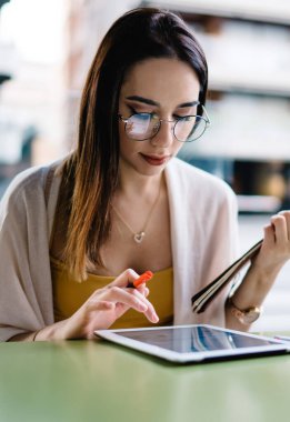 Gözlüklü ve yaz giysili konsantre bir kadın kampüs yakınlarındaki sokak kafede ödevlerini yaparken tablet kullanıyor ve notları kontrol ediyor.