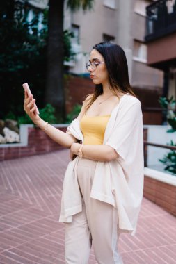 Gündüz vakti modern binaların önünde kendine güvenen akıllı telefon kullanan modaya uygun kıyafetli çekici bir kadın ekranına odaklanmış.