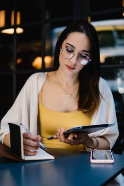 Gözlükleri ve günlük kıyafetleriyle konsantre olmuş genç bayan girişimci, tableti incelerken ve modern sokak kafesindeki masada otururken kalemle defterden not alıyor.