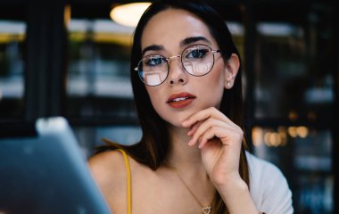 Ciddi genç girişimci gözlüklü ve günlük kıyafetleriyle tabletle çalışan modern açık hava kafeteryasında oturmuş kameraya bakan.